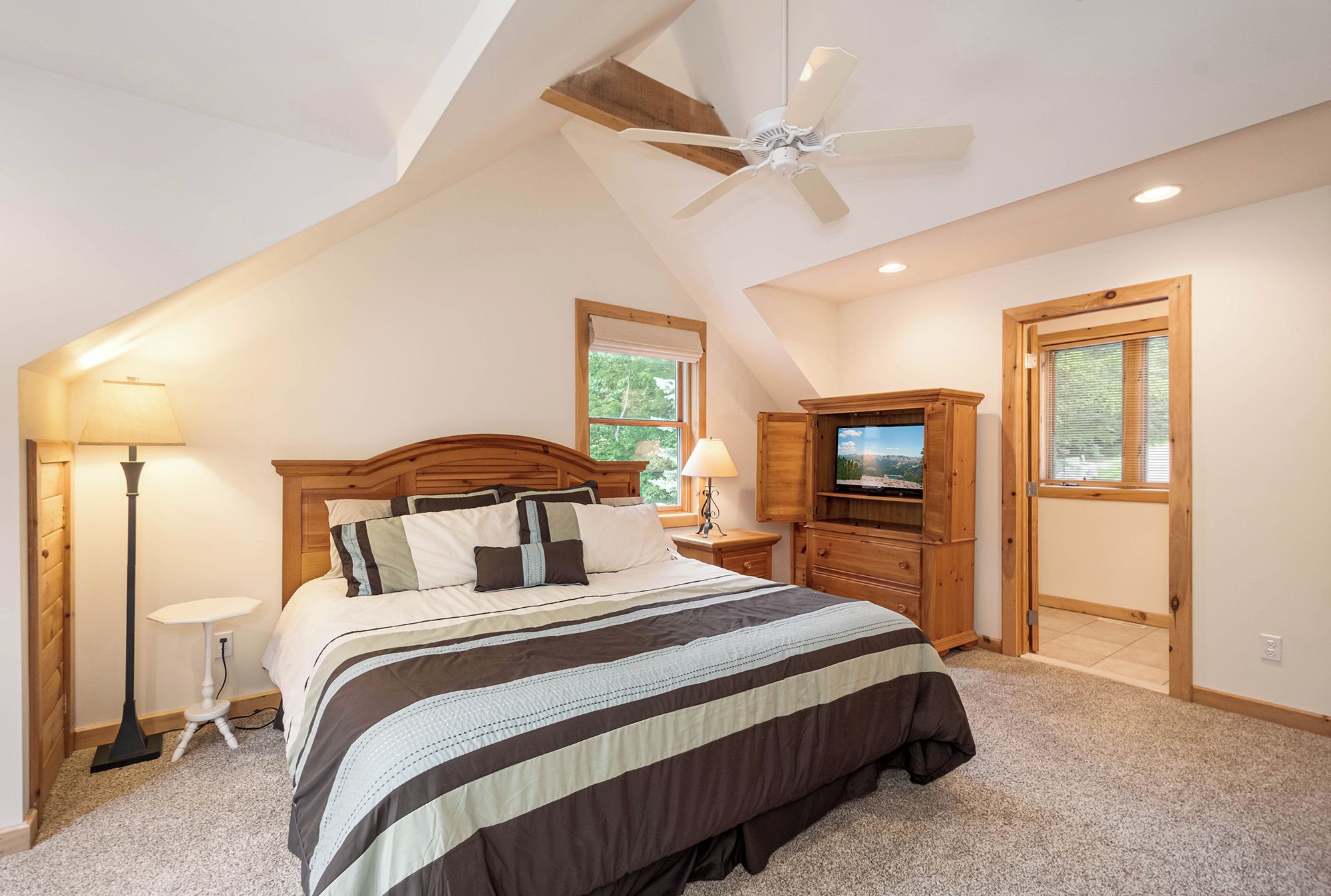 A bedroom with a large bed, wooden headboard, a ceiling fan, a TV, and a wooden cabinet.