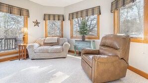 A cozy living room with a beige sectional sofa, a brown leather armchair, and a small table with a lamp. There are large windows with plaid valances and a star decoration on the wall. - Marsh Hill Villa by Taylor Made Deep Creek (McHenry)