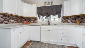 Une cuisine avec des meubles blancs, un pare-feu en carreaux, un évier et une fenêtre avec un rideau.