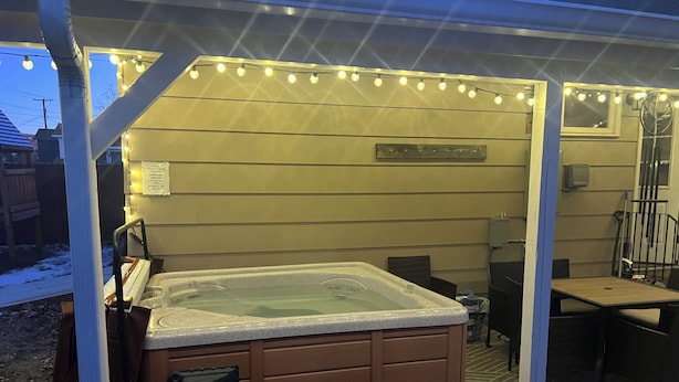 Un espace extérieur aménagé avec un jacuzzi, un banc en bois et une table, le tout illuminé par des guirlandes lumineuses.