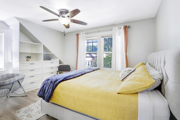 A bedroom with a bed, a ceiling fan, a window with curtains, a desk, and a chair.