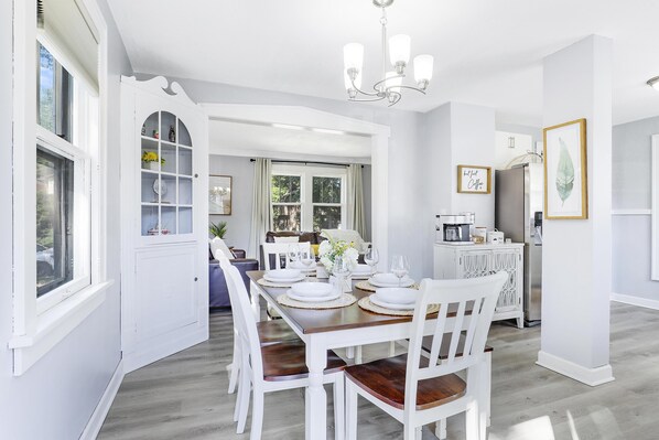 A dining area with a white table set for four, surrounded by white chairs, a chandelier, and a view of the outdoors through a window.