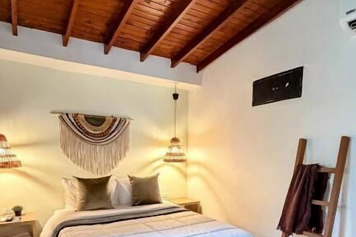 A bedroom with a wooden ceiling, a bed with a white bedspread, a hanging macrame wall decoration, and a wooden floor.
