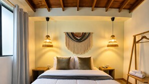 A bedroom with a large bed, two bedside tables, a wall hanging, and a wooden rack with towels. - Luum Guest House (Medellín)