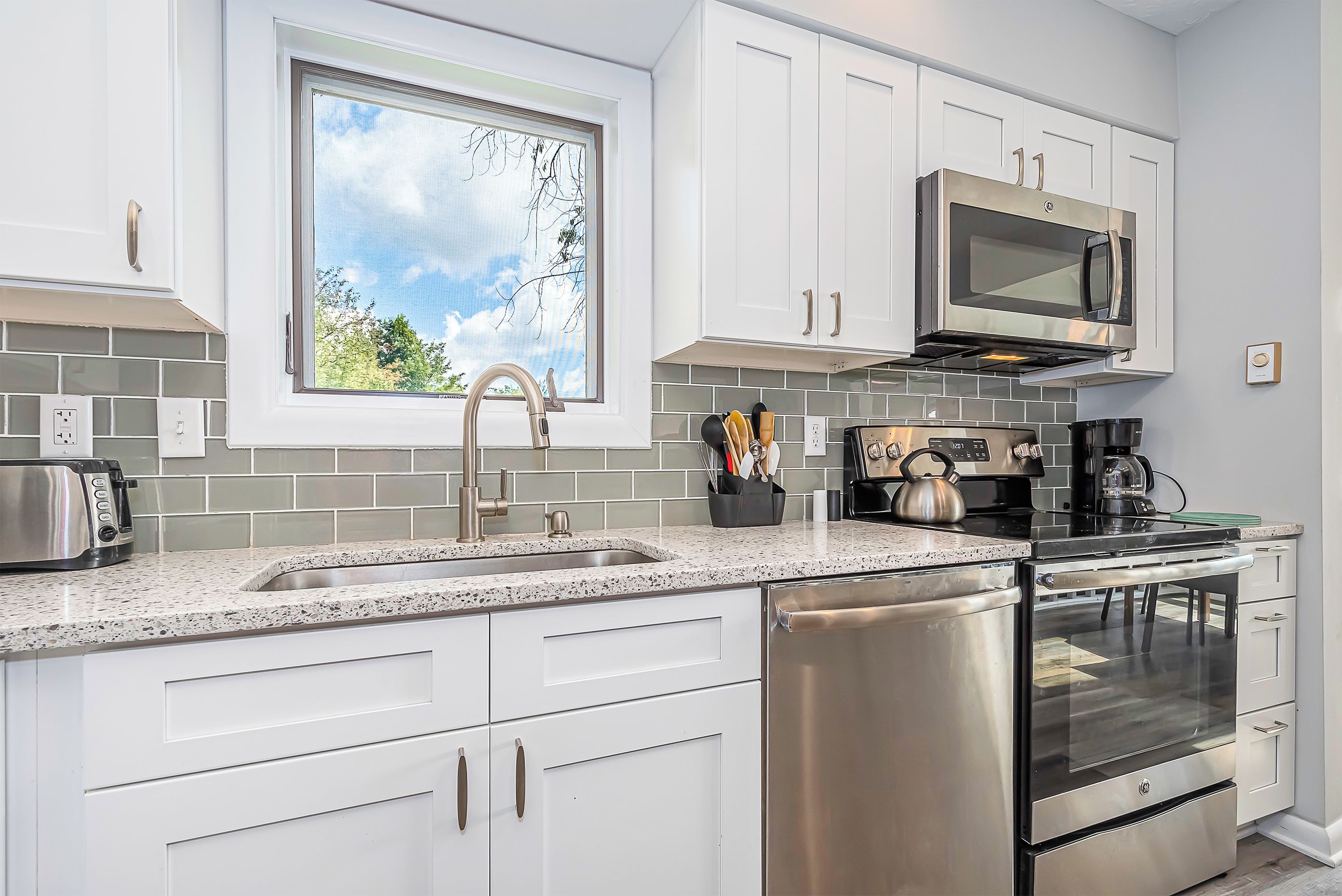 A modern kitchen with white cabinets, stainless steel appliances, a window with a view of trees, and a granite countertop.