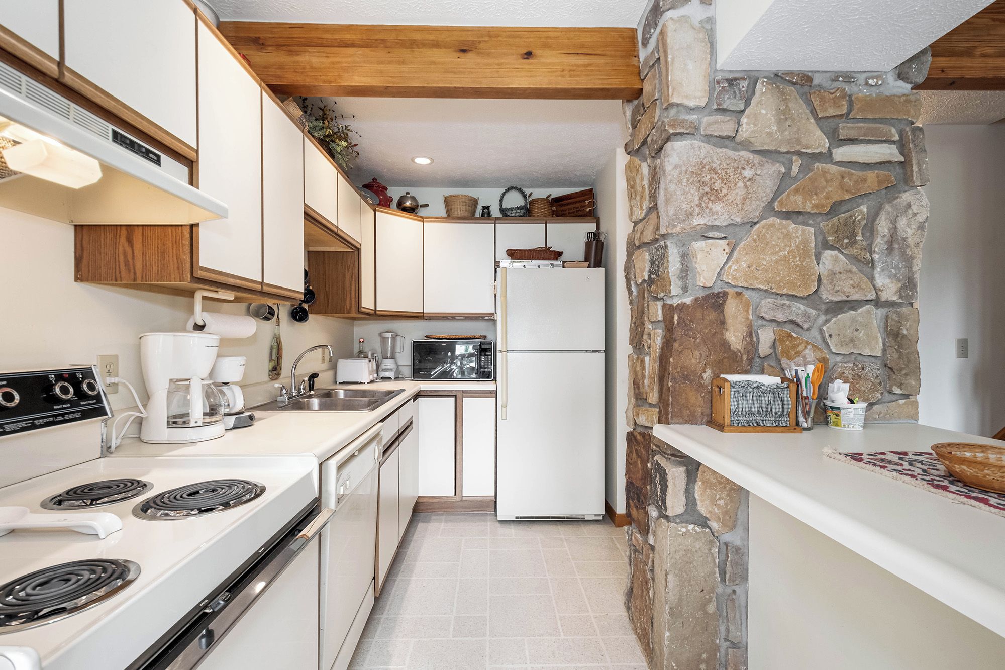 A compact kitchen with white appliances, wooden beams, and a stone wall.
