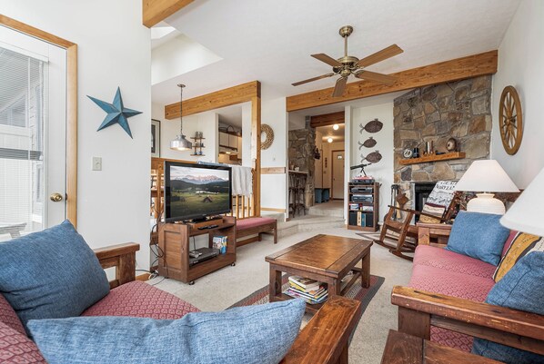 A cozy living room with a television, wooden furniture, a stone fireplace, and a ceiling fan.