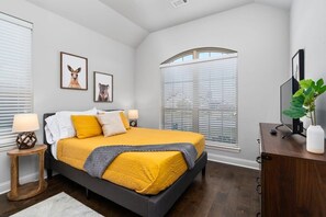 Une chambre à coucher avec un lit, deux tableaux encadrés, une fenêtre avec des stores, une table de chevet avec une lampe et une plante posée sur une commode.