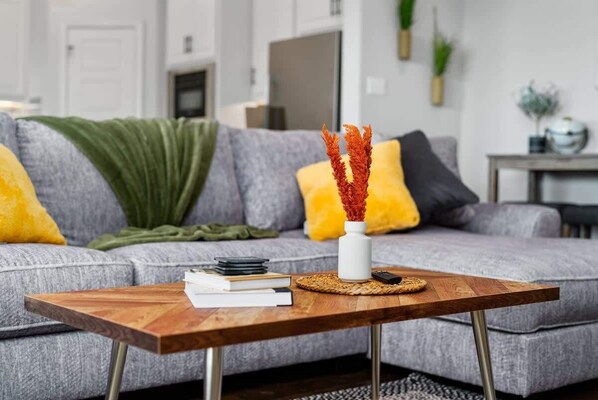 Un salon moderne avec un canapé gris, des coussins jaunes, une table basse en bois agrémentée de livres et d’un vase, et un plaid vert.