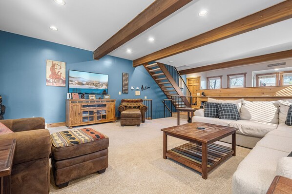 Una sala de estar acogedora con una pared azul, vigas de madera, una escalera y un televisor grande.