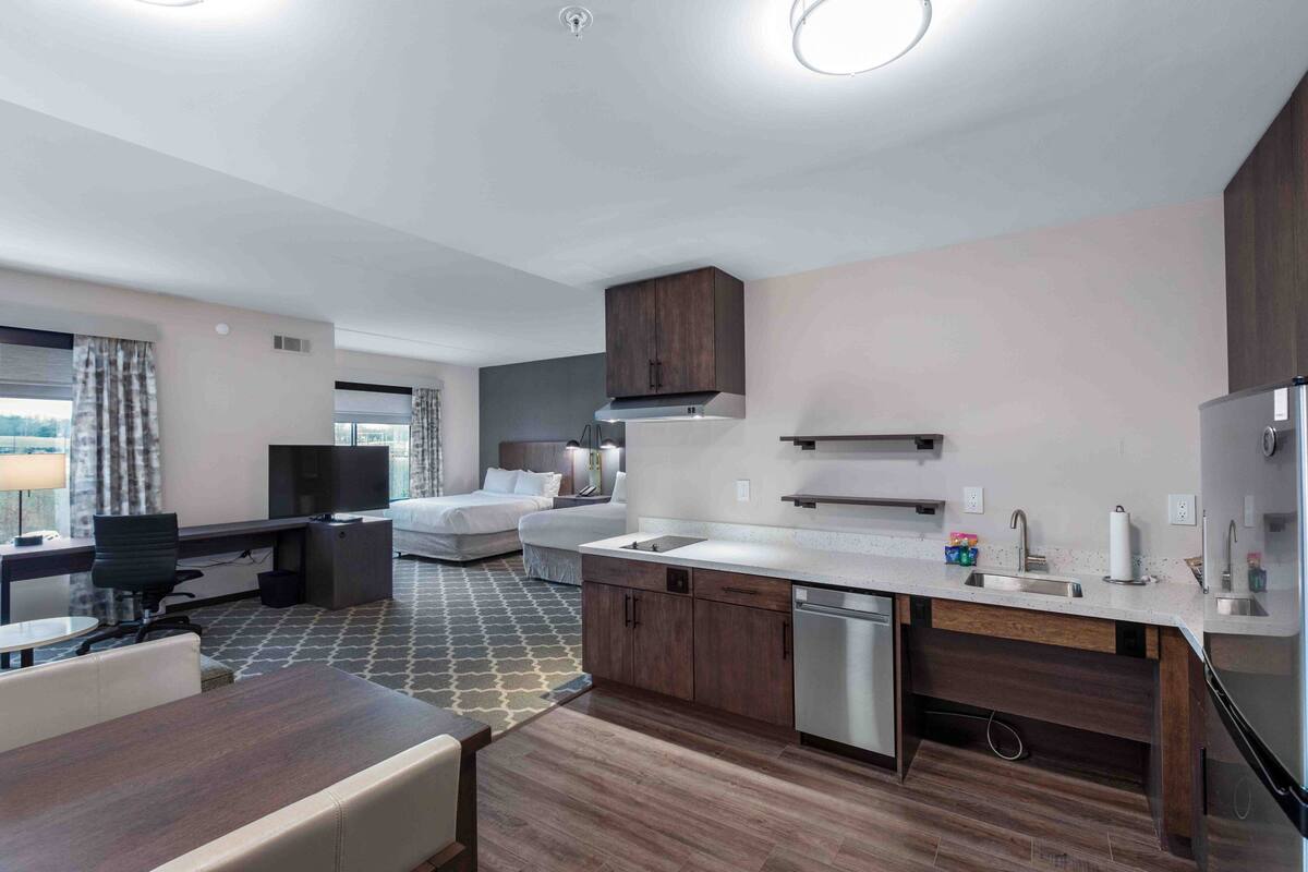 A modern hotel room with a kitchenette, a bed, a desk with a chair, and a TV.