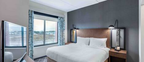 A hotel room with a large bed, a desk with a laptop, a nightstand with a lamp, and a view of the city through the window.