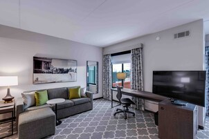 Una habitación de hotel moderna con un sofá gris, un escritorio con una silla, un televisor de pantalla plana, una lámpara y un ventanal grande con cortinas.