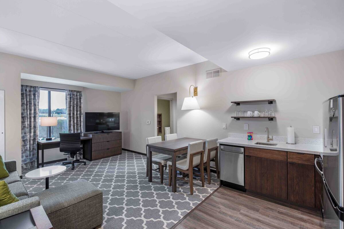 A hotel room with a living area, dining table, sofa, TV, kitchen, and a view of the city.