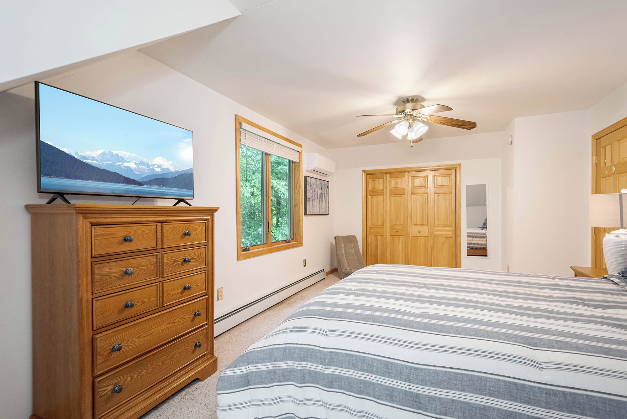 A bedroom with a bed, a dresser, a television, a ceiling fan, and a window with a view of trees.