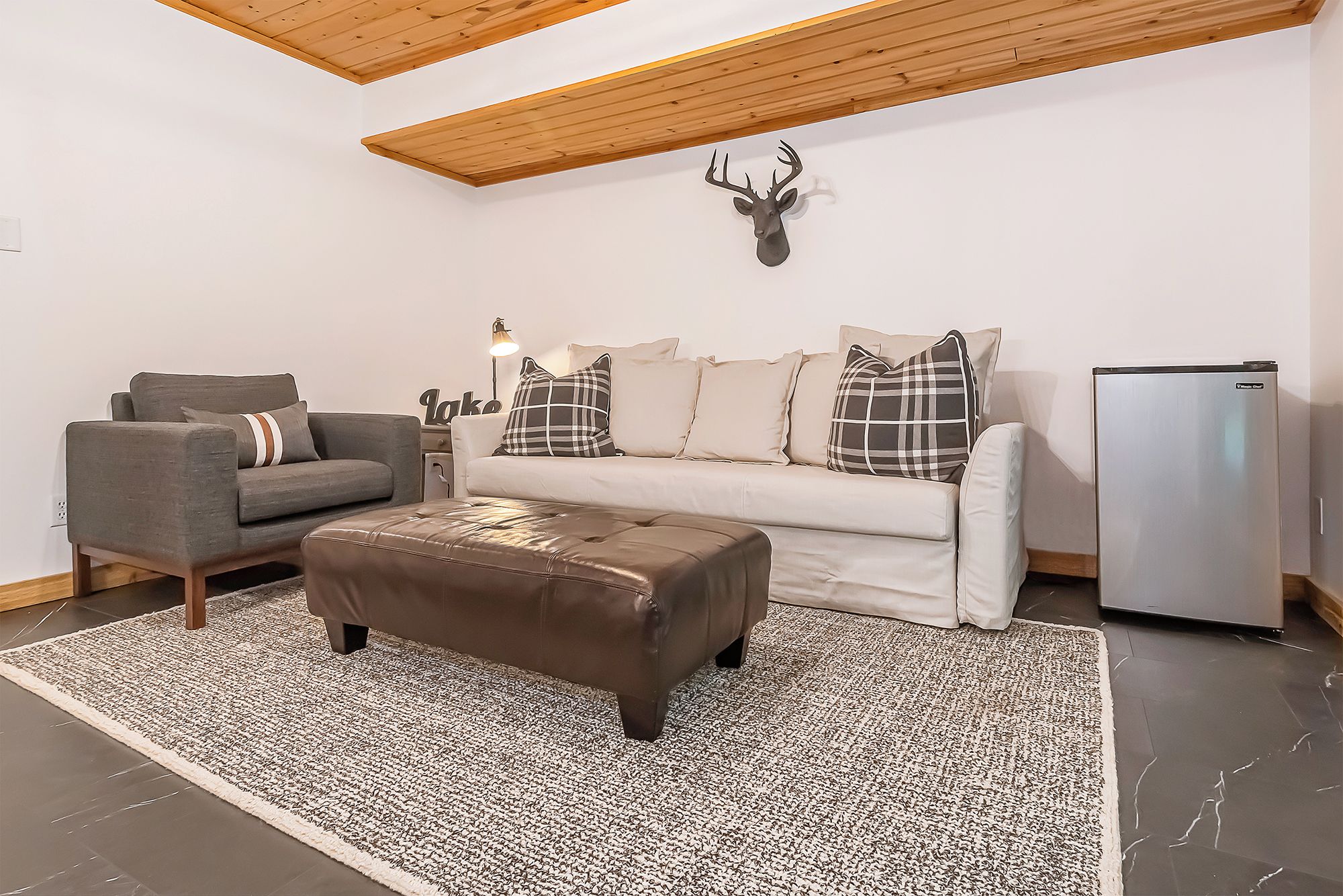 A living room with a grey sofa, a brown ottoman, a white sofa with plaid pillows, a mounted deer head, and a white refrigerator.