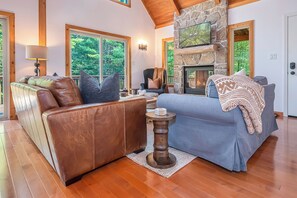 A spacious living room with a stone fireplace, leather sofas, and large windows offering a view of the outdoors. - Woodhaven by Taylor Made Deep Creek (Swanton)