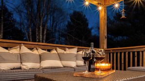 A wooden deck with a wicker sofa and cushions, a bottle of wine, and a candle on a wooden tray. - Woodhaven by Taylor Made Deep Creek (Swanton)