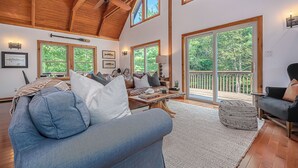A spacious living room with a high ceiling, large windows, and a view of the outdoors. - Woodhaven by Taylor Made Deep Creek (Swanton)