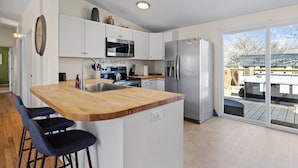 Cocina moderna con armarios blancos, una isla de madera con taburetes de bar, electrodomésticos de acero inoxidable y vistas a una terraza a través de puertas de cristal.