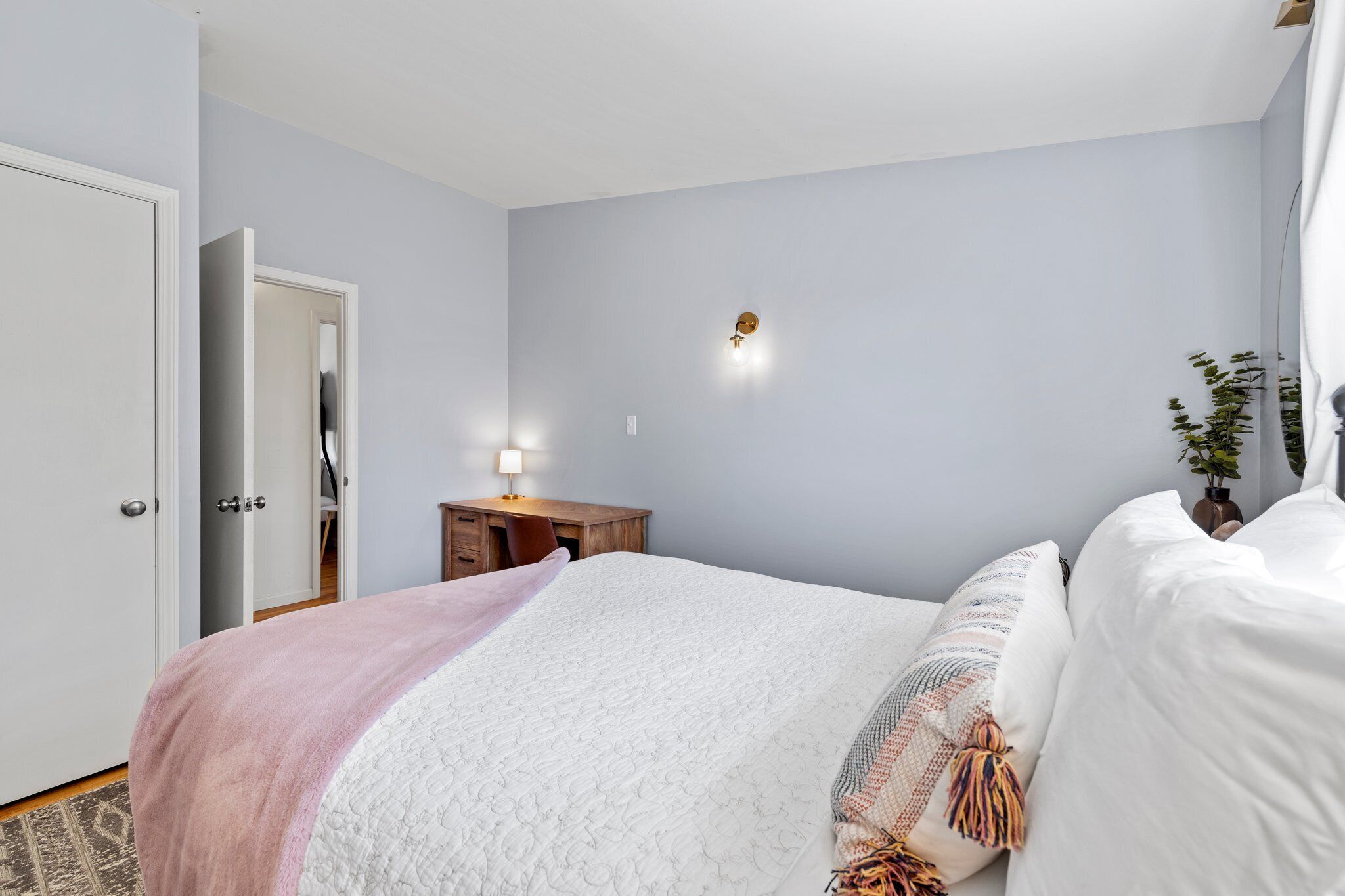 A bedroom with a bed, a bedside table with a lamp, a door, and a wall-mounted light fixture.