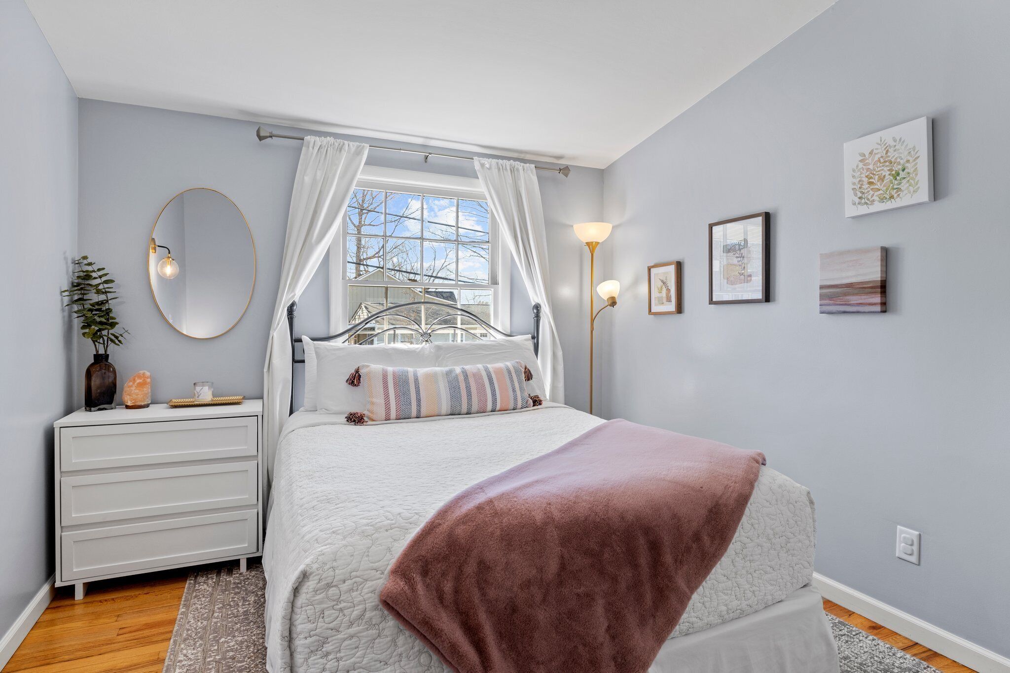 Un dormitorio con cama, mesita de noche, espejo, ventana con cortinas y una obra de arte en la pared.