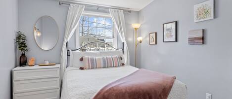 Un dormitorio con cama, mesita de noche, espejo, ventana con cortinas y una obra de arte en la pared.