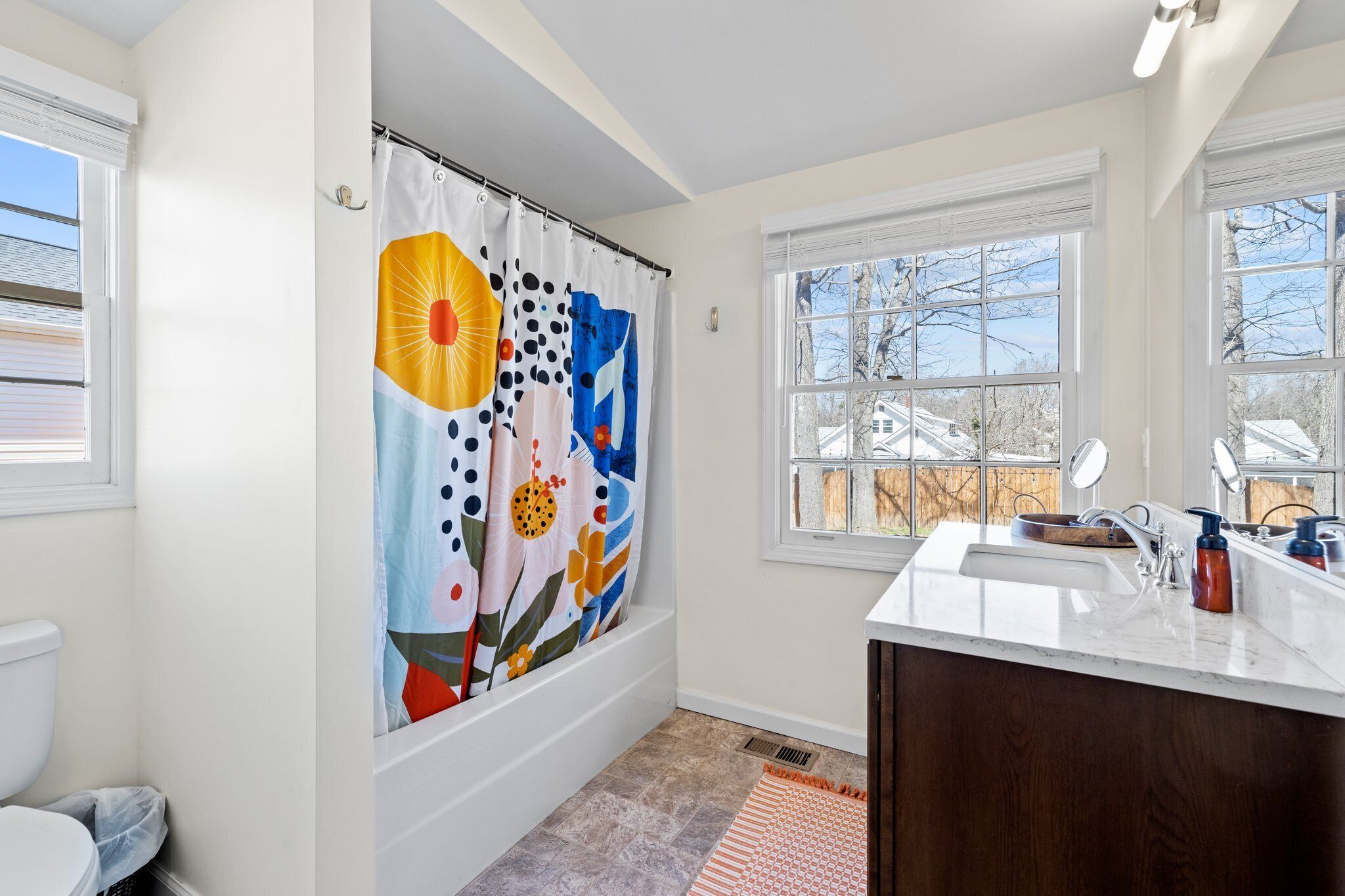 Une salle de bain avec un rideau de douche orné d’un motif floral coloré, un lavabo blanc avec un meuble-vasque en bois, et une fenêtre donnant sur les arbres et les bâtiments à l’extérieur.