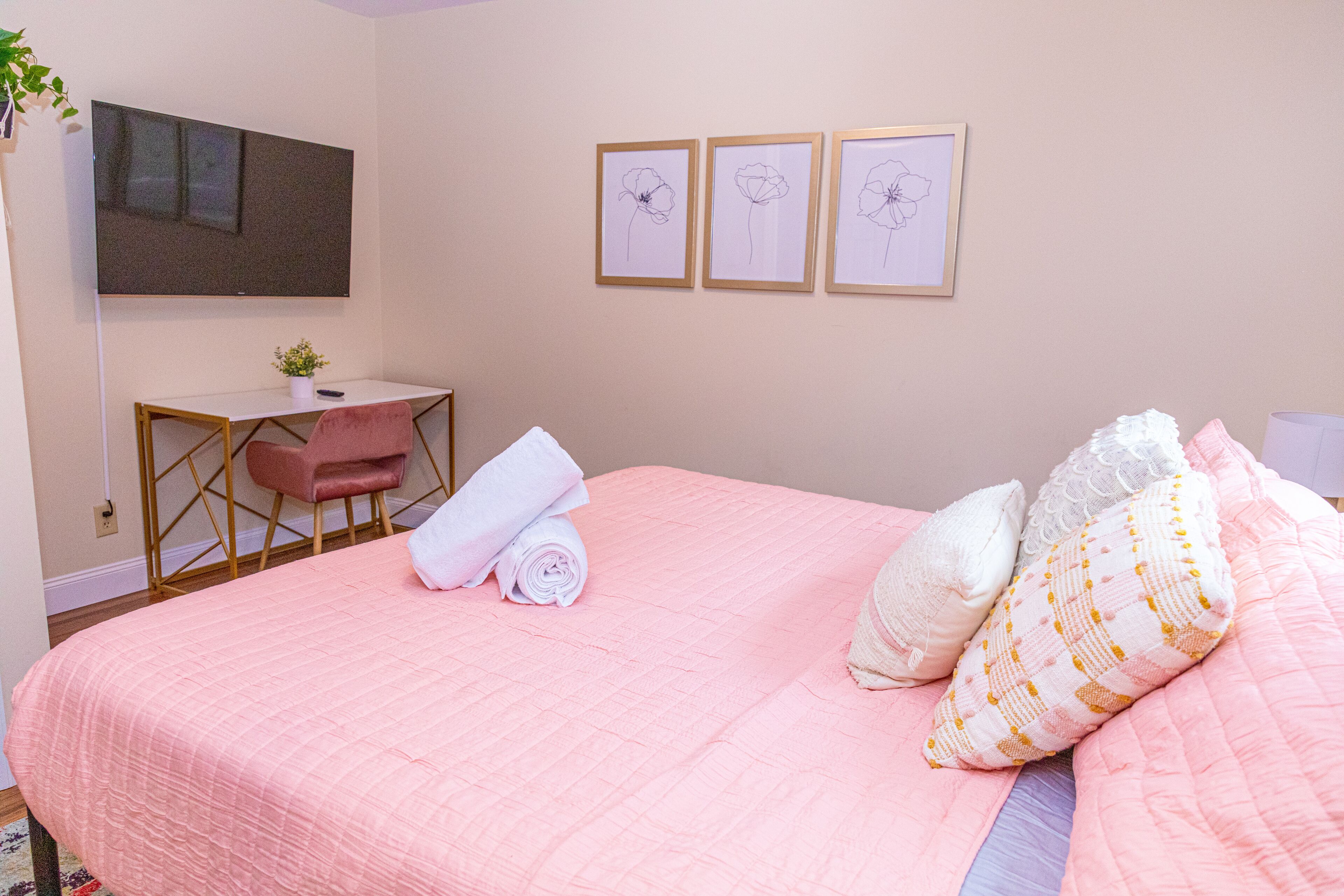 A bedroom with a pink bedspread, a desk with a chair, a flat-screen TV, and three framed floral pictures on the wall.