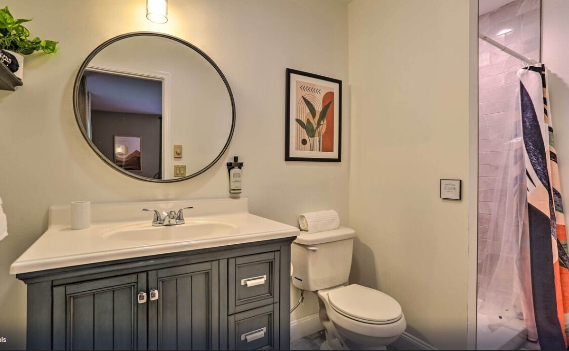 Une salle de bain avec un miroir rond, un lavabo muni d’un porte-serviette, des toilettes et un rideau de douche.