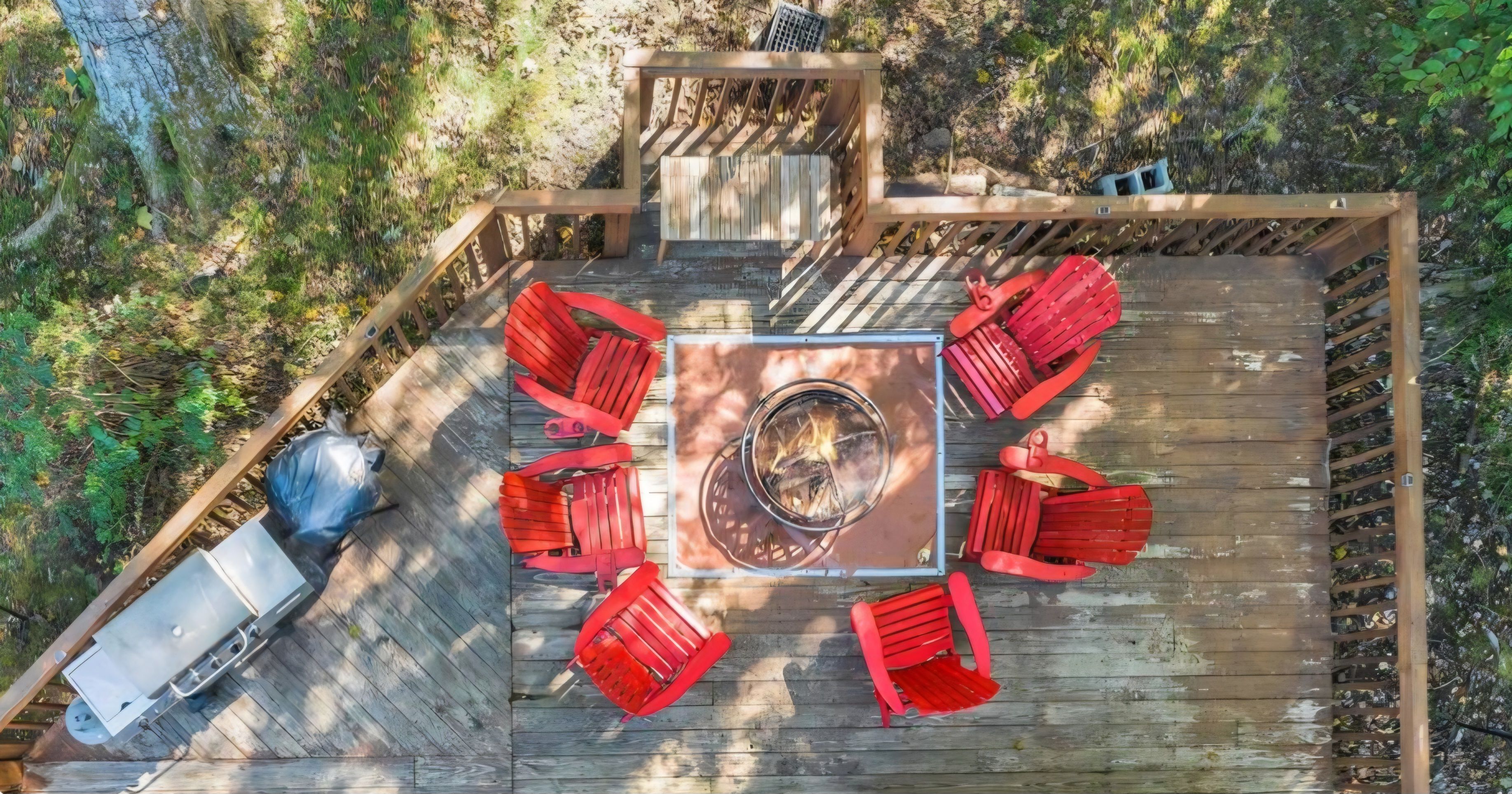 A deck with a central fire pit surrounded by red chairs, a table, and a bench.
