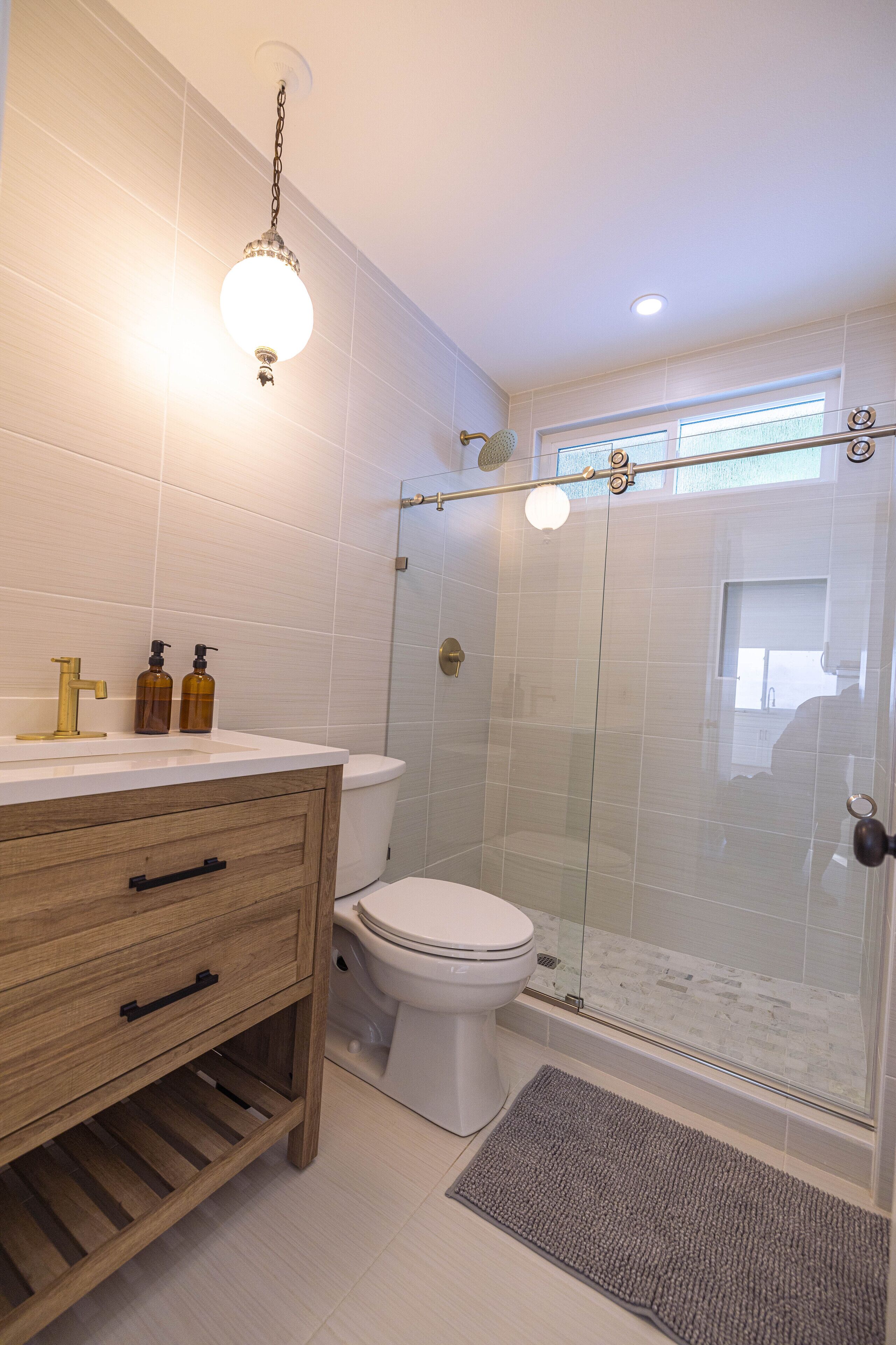 A bathroom with a wooden vanity, white tiles, a glass shower enclosure, and a visible window.