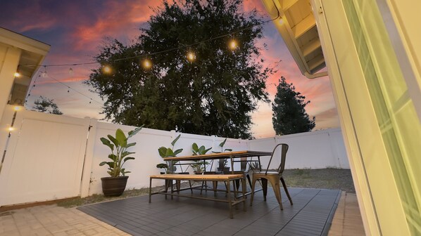 Un patio al aire libre con mesa y sillas de madera, rodeado de plantas en macetas y luces colgantes.