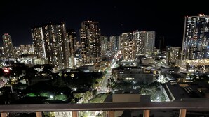 A balcony with a wooden table and a glass of amber liquid. In the background, a cityscape with illuminated buildings is visible. - Newly remodeled 2 bd/2ba Kaka'ako condo on 25th floor with city/ocean views (Honolulu)