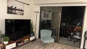 A modern living room with a flat-screen TV, a light blue armchair, and a sliding glass door leading to a balcony with a city view. - Newly remodeled 2 bd/2ba Kaka'ako condo on 25th floor with city/ocean views (Honolulu)