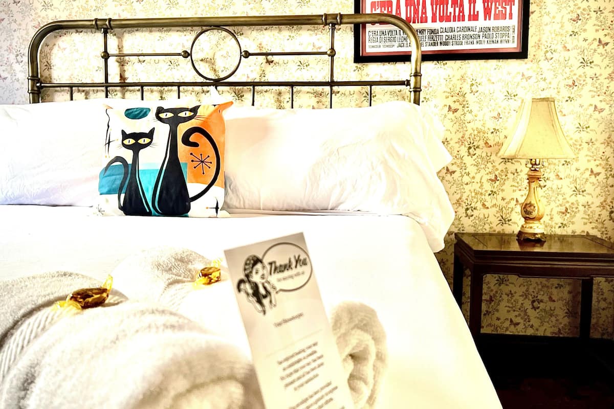 A neatly made bed with a floral-patterned headboard, a bedside table with a lamp, and a framed menu on the wall.