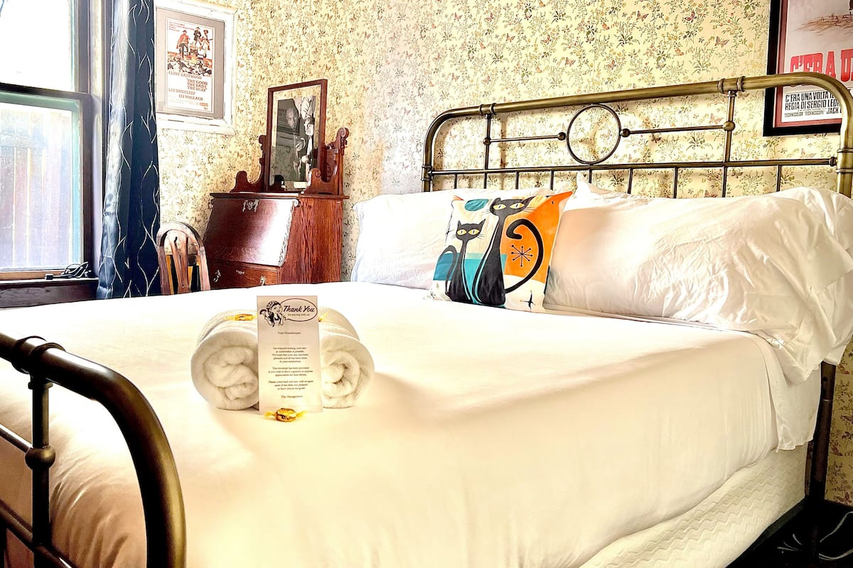 A neatly made bed with a folded towel and a thank you note, a metal bed frame, a window with blinds, and floral wallpaper.