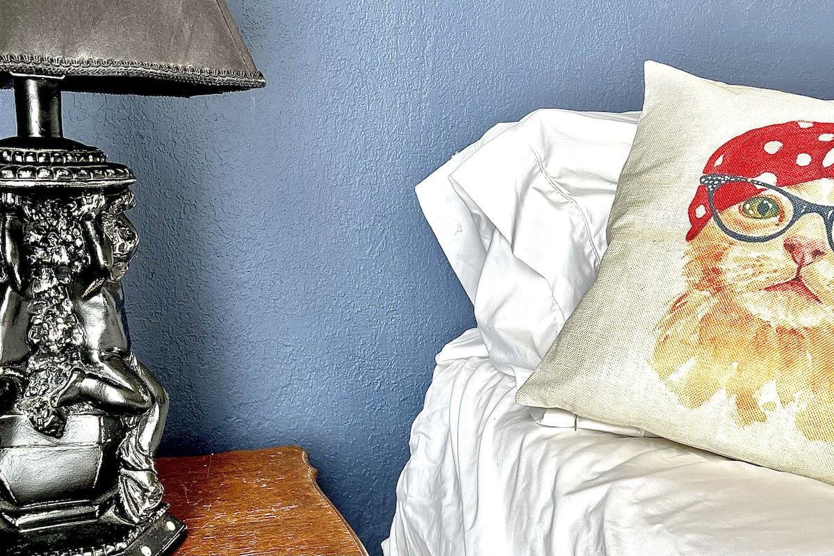 A bed with white bedding and a pillow featuring a red polka-dotted hat and glasses.