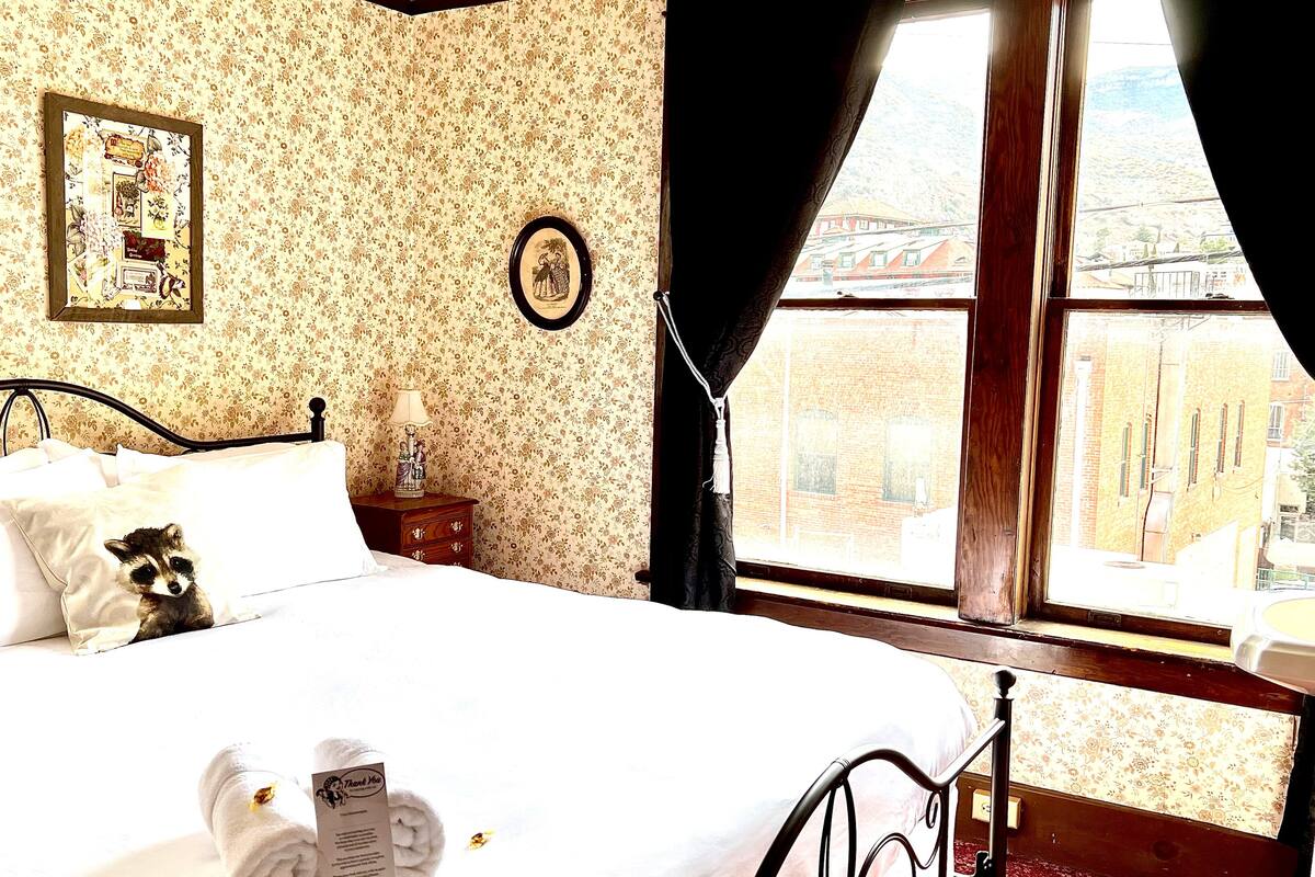 A bedroom with a bed, a nightstand, a lamp, a framed picture, and a window with a view of buildings.