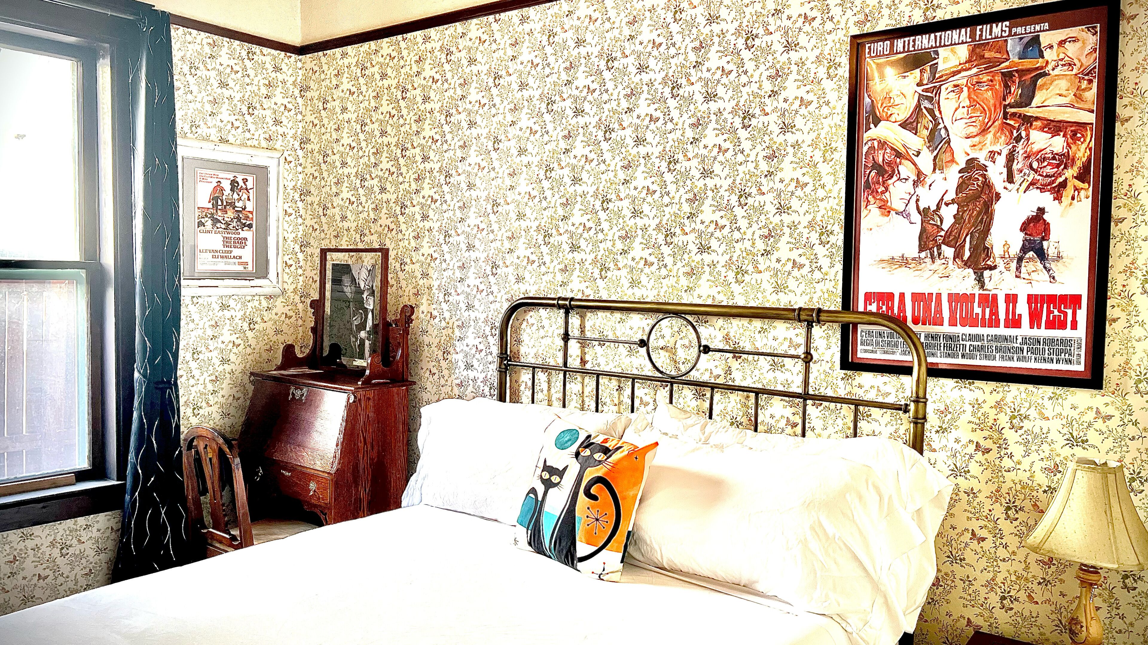 A bedroom with floral wallpaper, a metal bed frame, a poster on the wall, and a window with curtains.