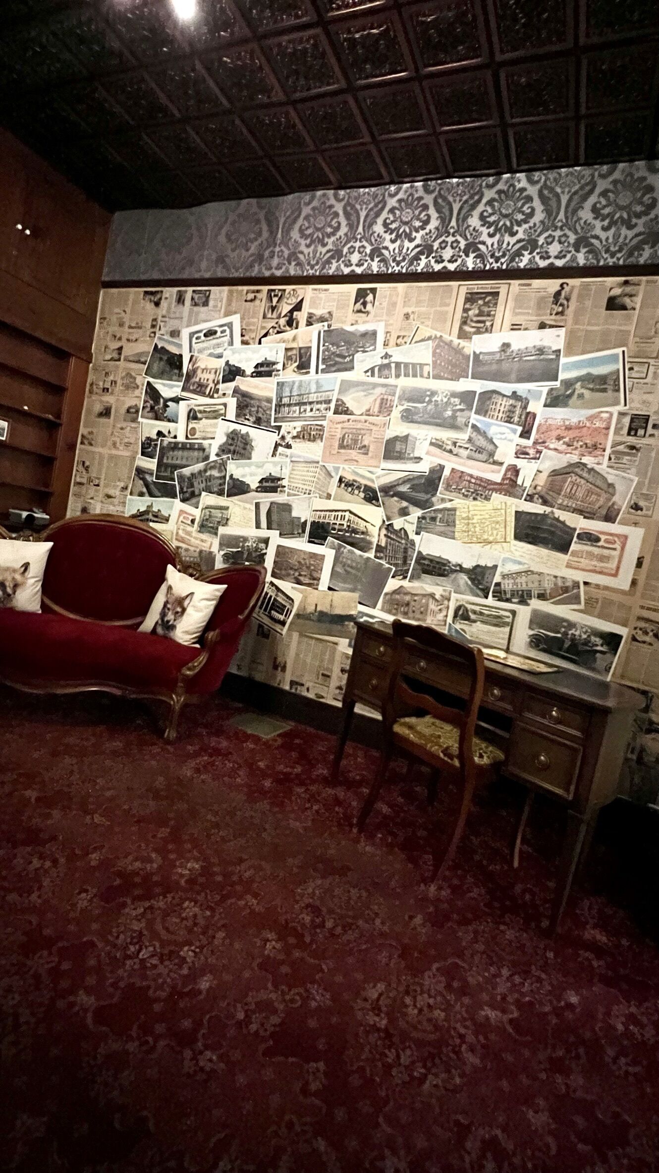 Ein Zimmer mit einem roten Sofa, einem Holzstuhl und einer Wand, die mit verschiedenen Fotos und Zeitungsartikeln bedeckt ist.