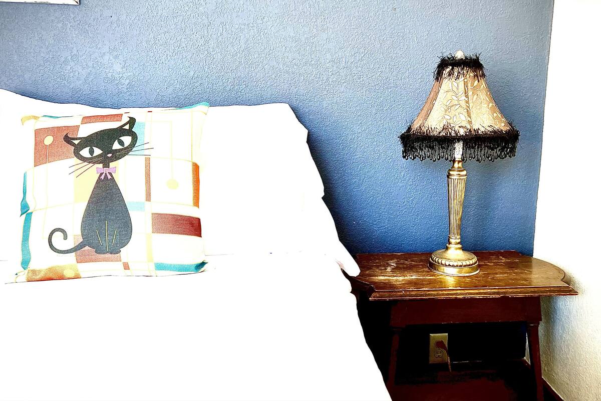 A room with a blue wall, a lamp on a wooden side table, and a partially visible bed with a pillow featuring a cat design.