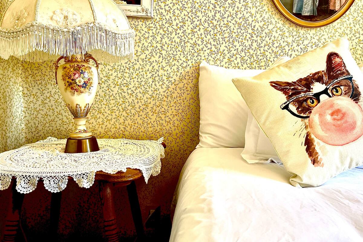A bed with a pillow featuring a cat illustration, a bedside table with a lamp, and a mirror on the wall.