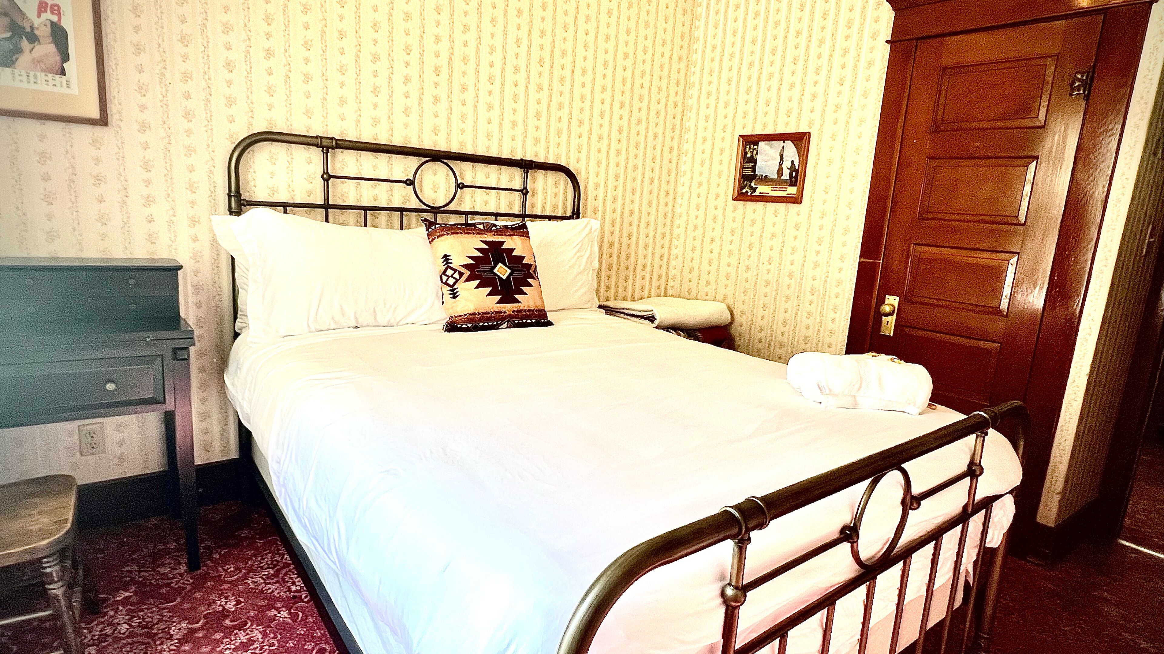 Ein Einzelbett mit Metallgestell, weißer Bettwäsche und einem Zierkissen. Eine Holztür und ein kleines gerahmtes Bild sind an der Wand zu sehen.
