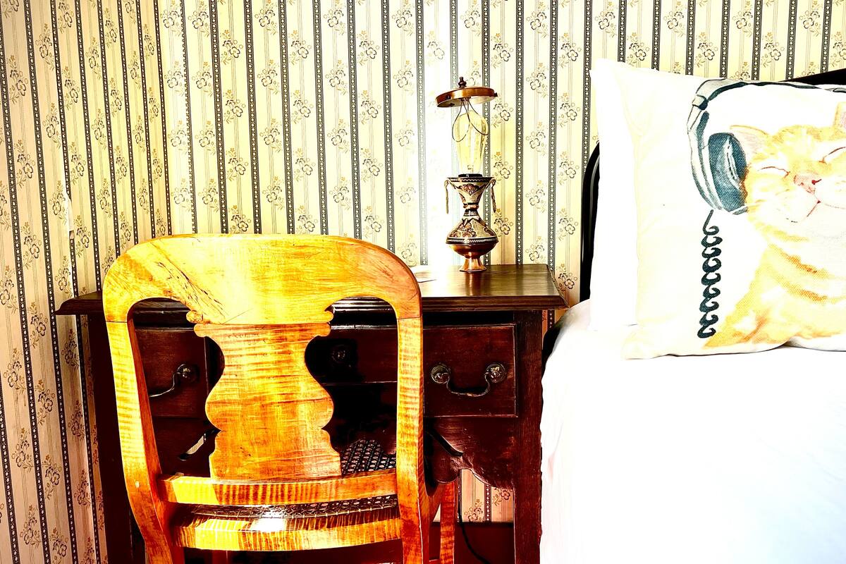 A wooden chair with a unique backrest design, a bedside table with a lamp, and a bed with a pillow featuring a cat illustration.