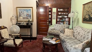 Un salon chaleureux avec un canapé, un fauteuil et une petite table sur laquelle sont posés des livres. On y trouve une porte en bois, une bibliothèque et une lampe.