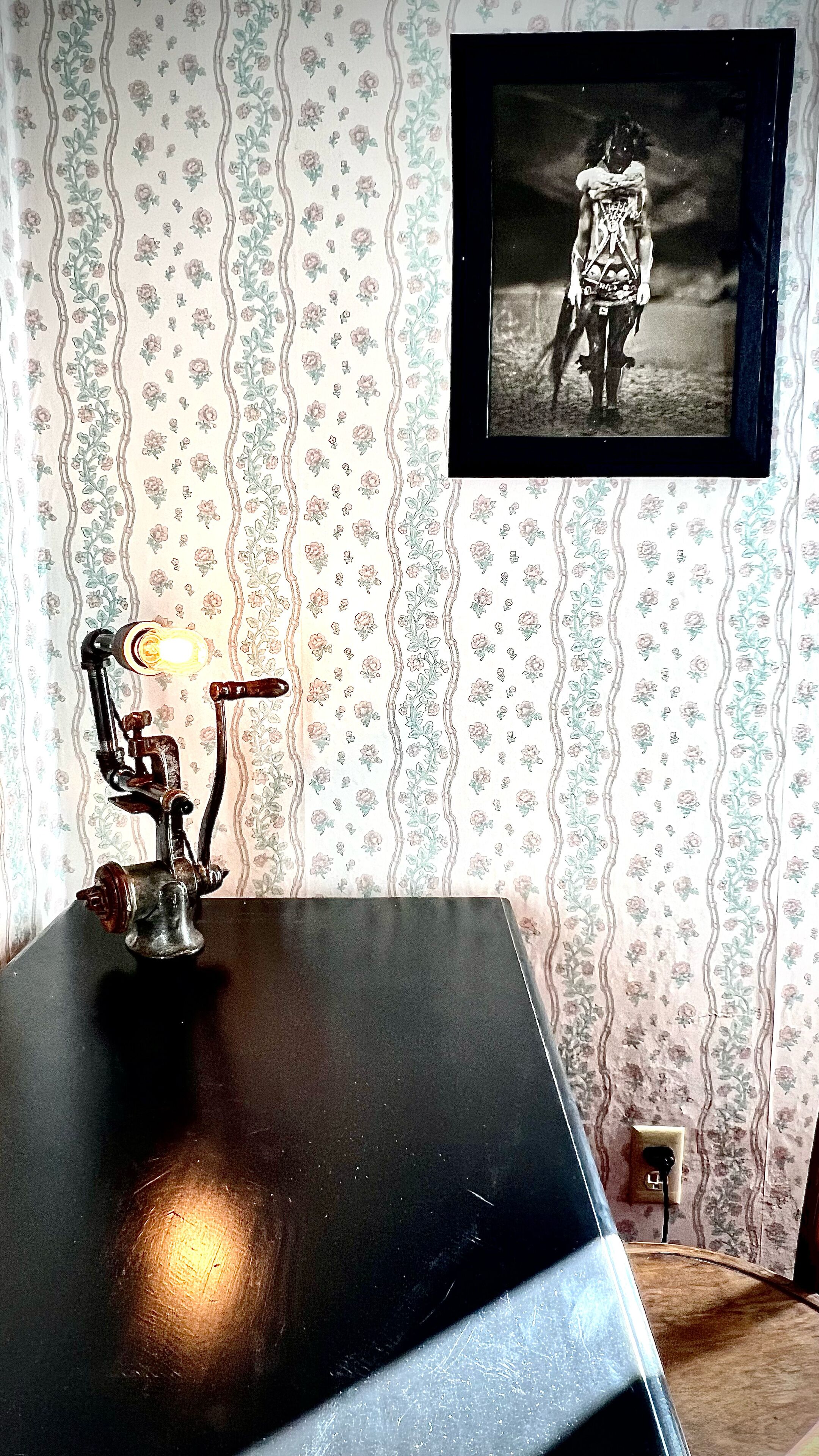 A room with a black table, a vintage lamp, floral wallpaper, and a framed photograph on the wall.