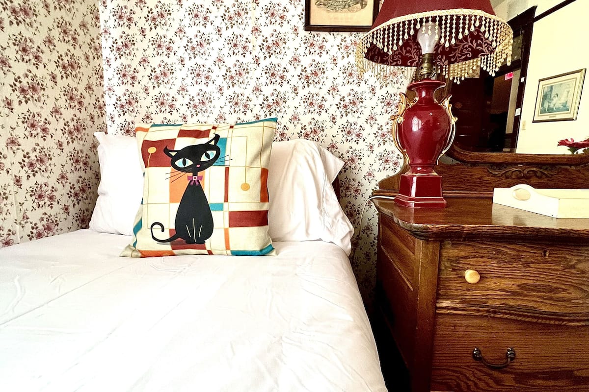 A bedroom with floral wallpaper, a bed with a checkered pillow, a wooden dresser with a mirror, and a red lamp.
