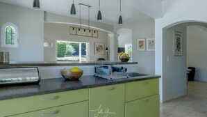 A kitchen with green cabinets, a large window, pendant lights, and a bowl of fruit on the counter.