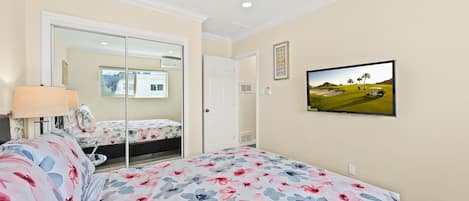 Quarto com cama, espelho, televisão na parede e porta que dá para outro espaço.